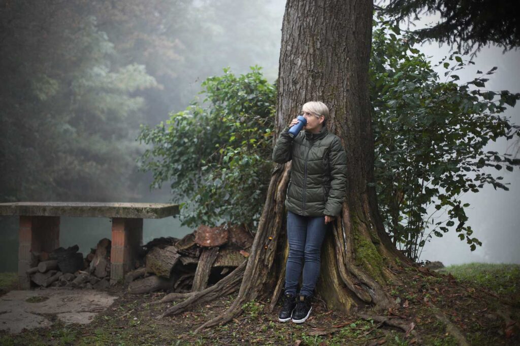 Žena u zelenoj jakni oslonjena na drvo u maglovitoj šumi, pije iz termosice, okružena prirodom.