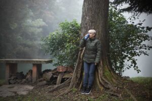 Žena u zelenoj jakni oslonjena na drvo u maglovitoj šumi, pije iz termosice, okružena prirodom.